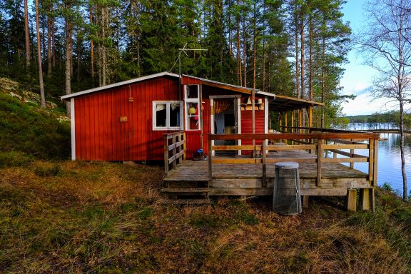 Röd stuga vid en sjö omgiven av träd och grönska.