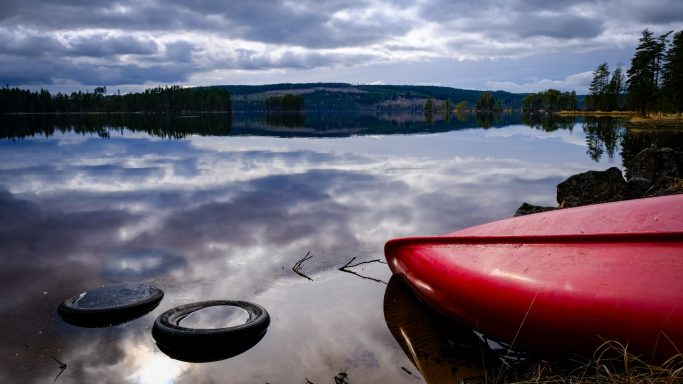 Röd kajak vid en spegelblanka sjö med molnig himmel och skog i bakgrunden.
