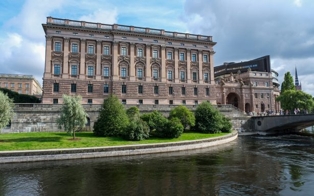 Svenska riksdagshuset med grönskande omgivningar och vattendrag i förgrunden.